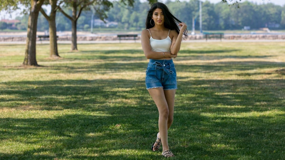 Young woman standing confidently in a park, wearing denim shorts and a crop top.