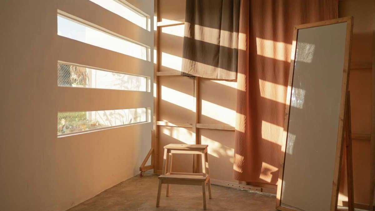 A minimalist room in Indonesia with warm sunlight, featuring a wooden stool, mirror, and curtains.