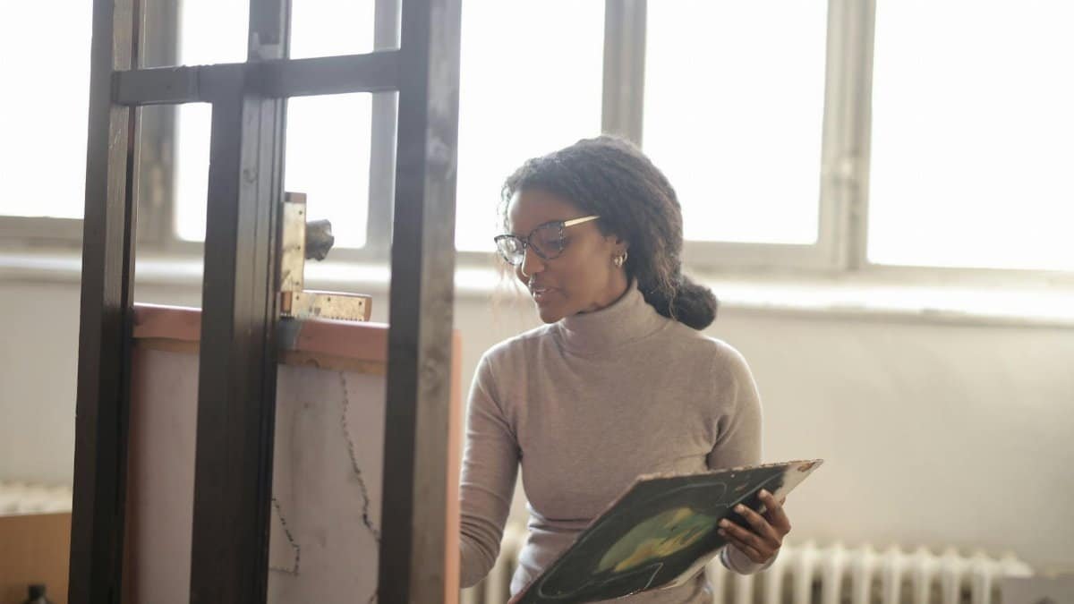 Positive young African American lady in beige turtleneck and eyeglasses standing near easel against bright window and drawing picture in creative studio
