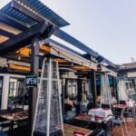 Sunny outdoor restaurant setting with tables, chairs, and heaters under a modern pergola.