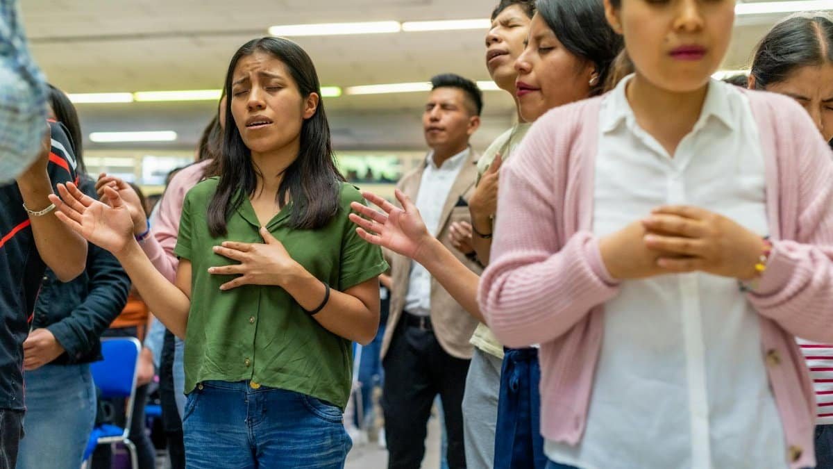 A diverse group of people standing indoors, engaging in collective prayer with closed eyes and devotional expressions.