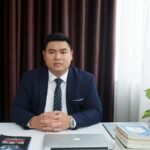 Businessman in a formal suit sitting at a desk with a laptop and books in a modern office setting.