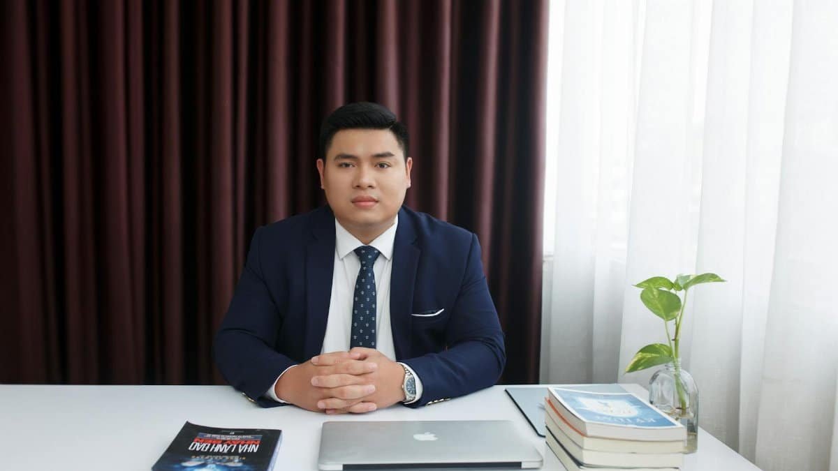 Businessman in a formal suit sitting at a desk with a laptop and books in a modern office setting.