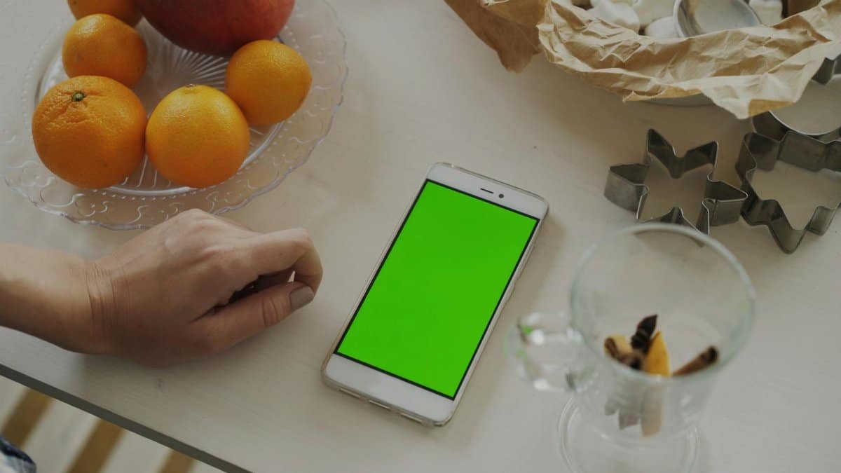 Top view of a smartphone with a green screen next to a plate of oranges and an apple on a table.