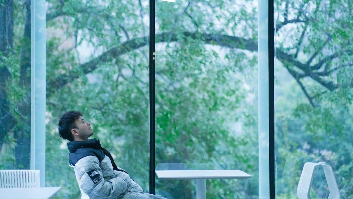 Man sitting indoors by large glass windows with lush greenery outside, creating a serene atmosphere.