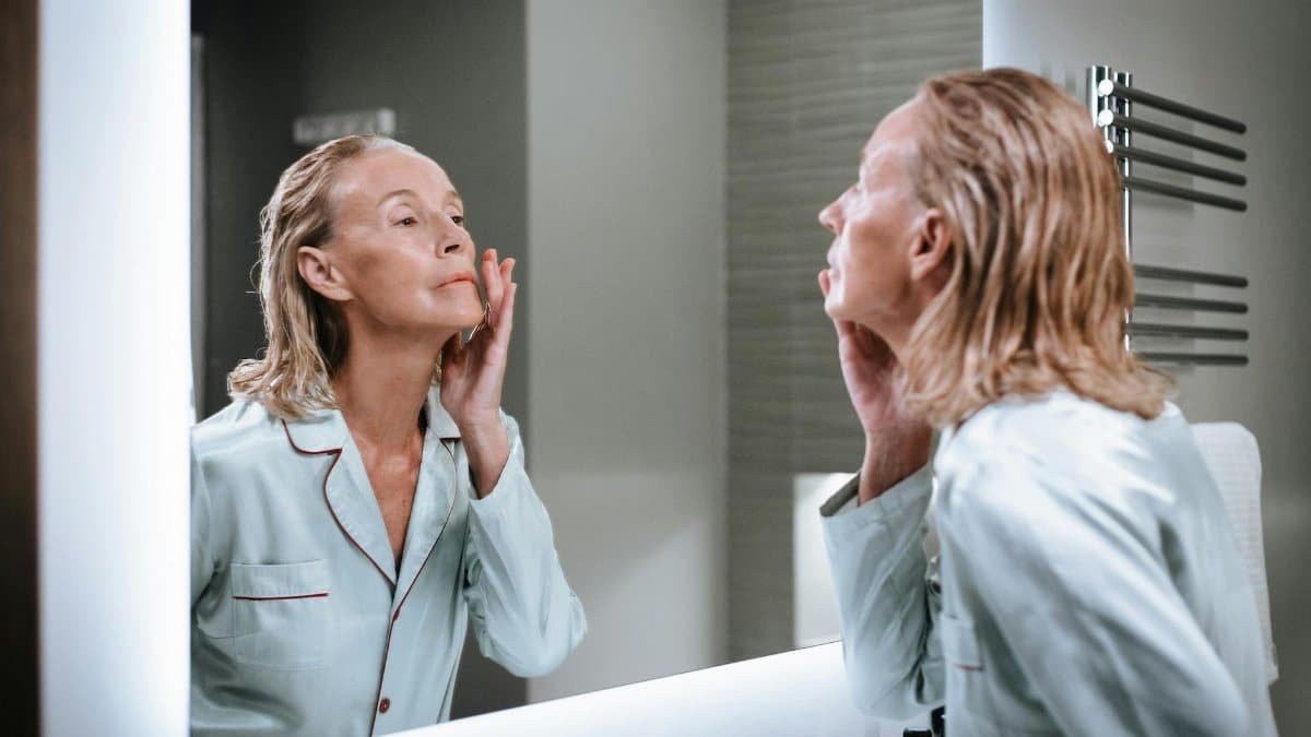 Elderly woman admiring her skincare results in a well-lit bathroom mirror.