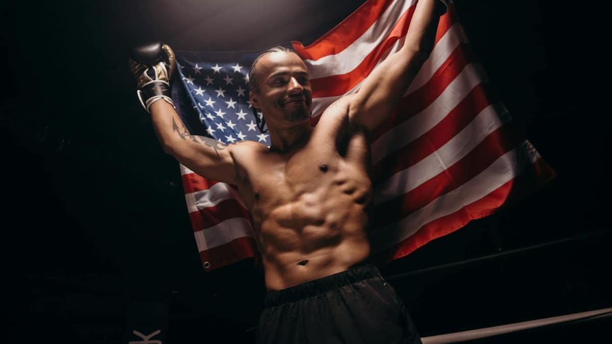 Muscular male boxer celebrates victory with American flag in the boxing ring.
