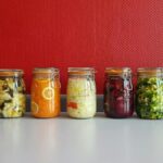 A row of vibrant, multicolored mason jars filled with assorted fermented vegetables against a red wall.