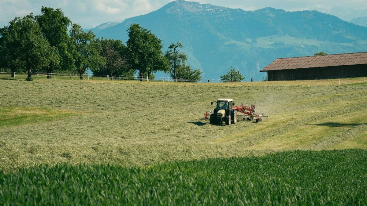 A tractor rakes hay in a scenic mountain farm setting on a sunny day, surrounded by lush fields and trees.