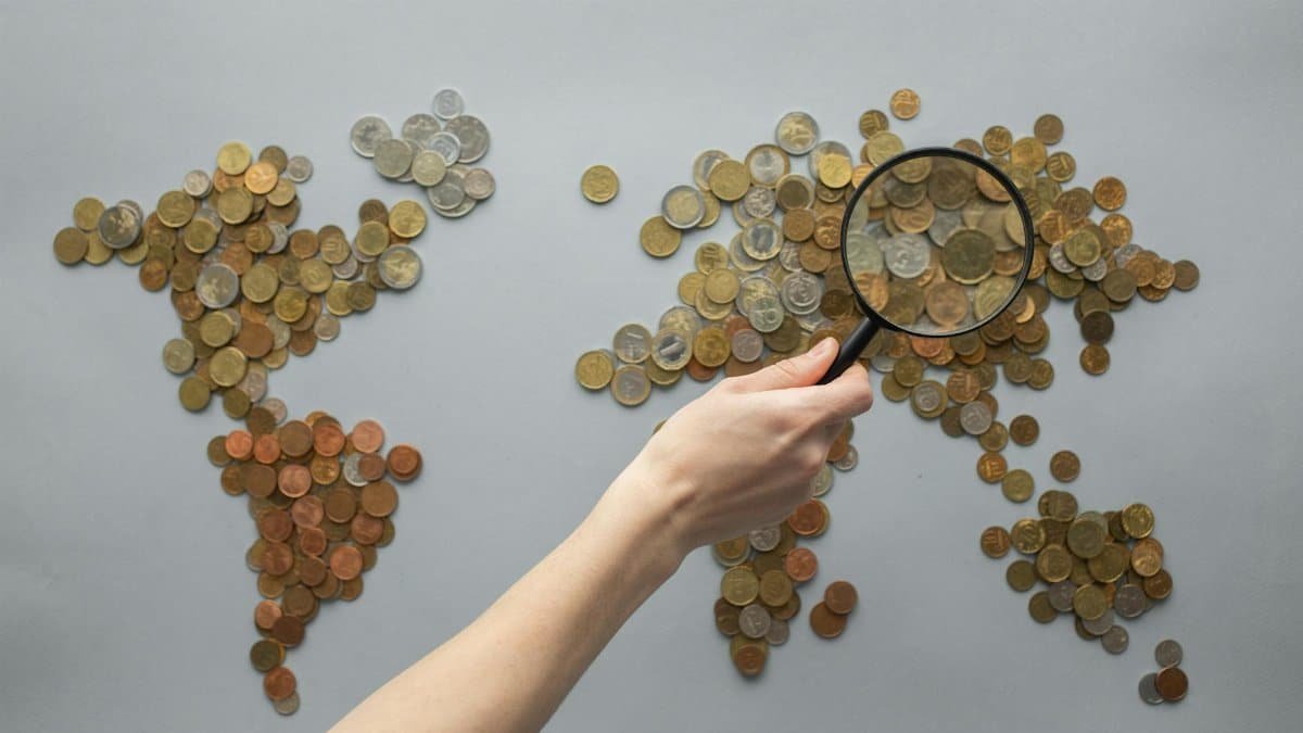 Top view of crop unrecognizable traveler with magnifying glass standing over world map made of various coins on gray background