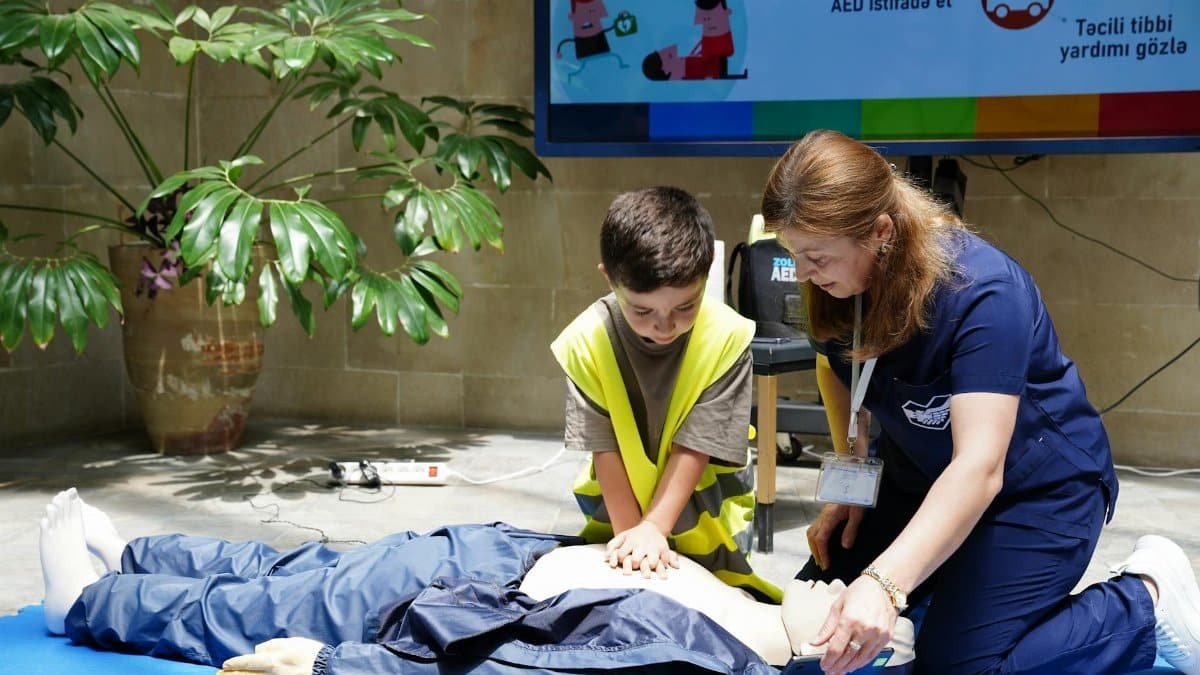 A young boy learning CPR from an instructor on a dummy indoors, with educational materials displayed.