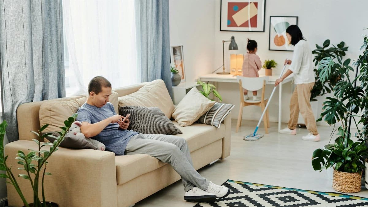 A family engages in daily chores, with a man relaxing and a woman mopping the floor.