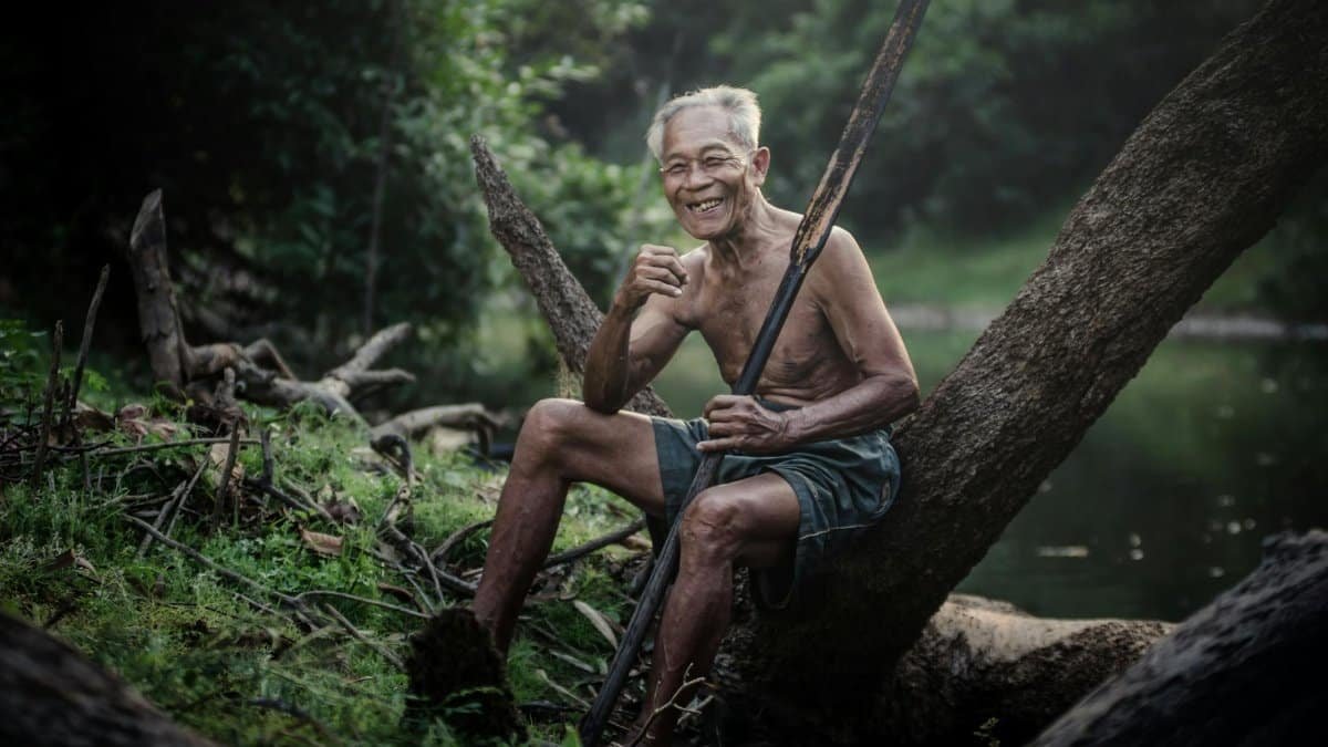 A senior man smiles happily while sitting on a log by a tranquil river, embodying peace.