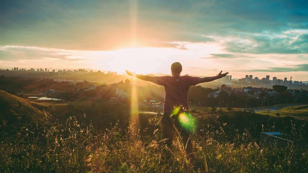 Person standing on grassy hill with open arms embracing a vibrant sunset over a cityscape.