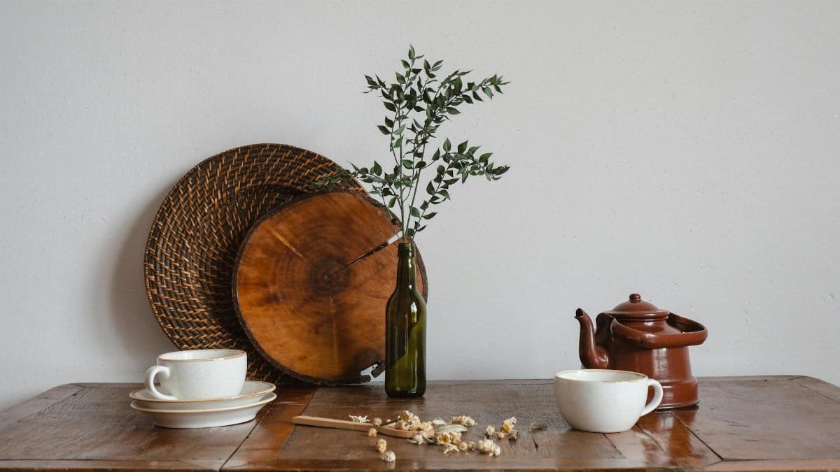 A rustic still life showcasing a teapot, cups, and a bottle with branches on a wooden table.
