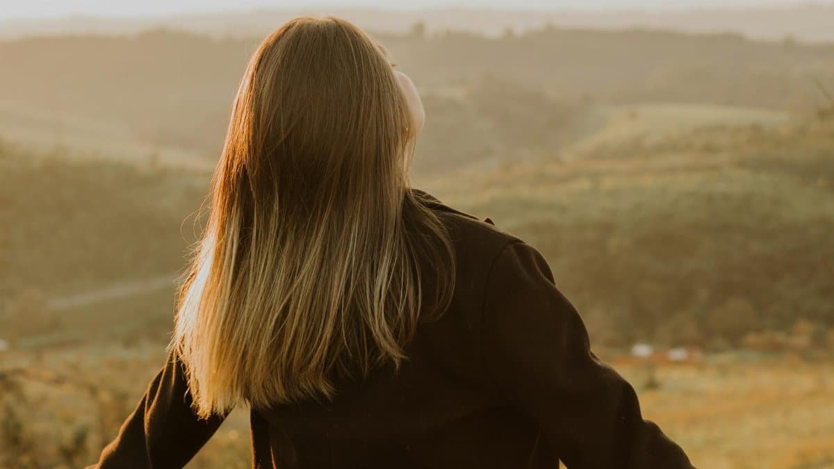 Back view of a woman with long hair standing in a scenic hillside at sunset, embracing nature.