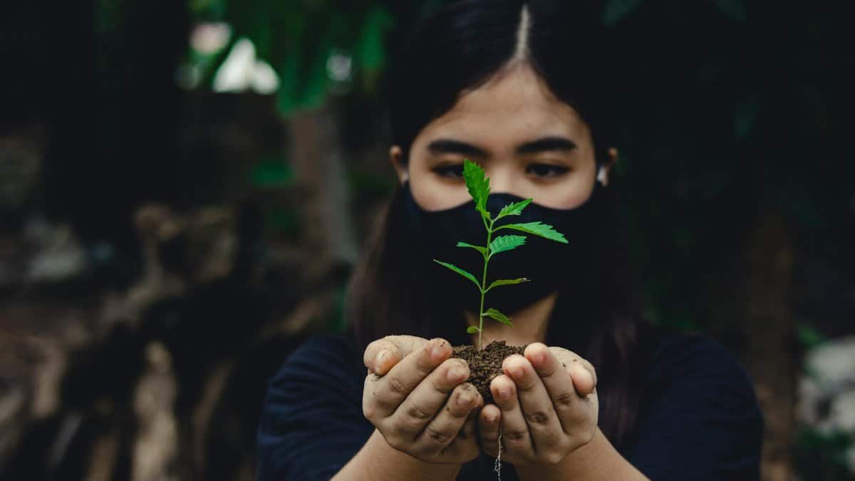 Woman in mask holds a small plant with soil, symbolizing environmental care.