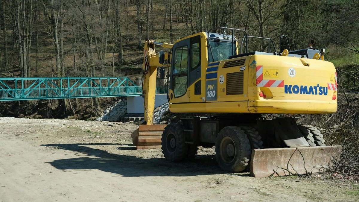 Yellow excavator parked near a blue bridge at a construction site in early spring.