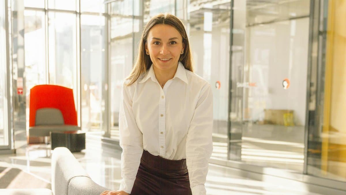 A confident woman in a white shirt stands in a modern office, radiating professionalism and success.