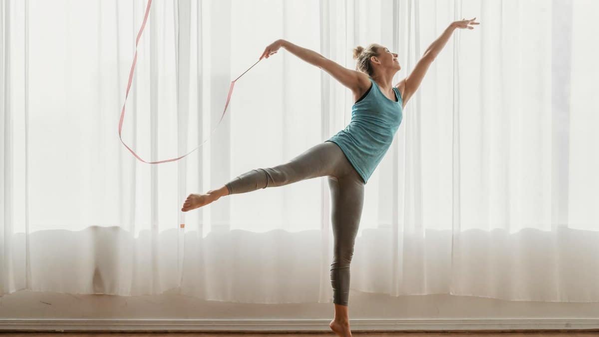 Full body of barefoot young female athlete performing dance in swallow pose with gymnastic equipment in front of window with light white curtains