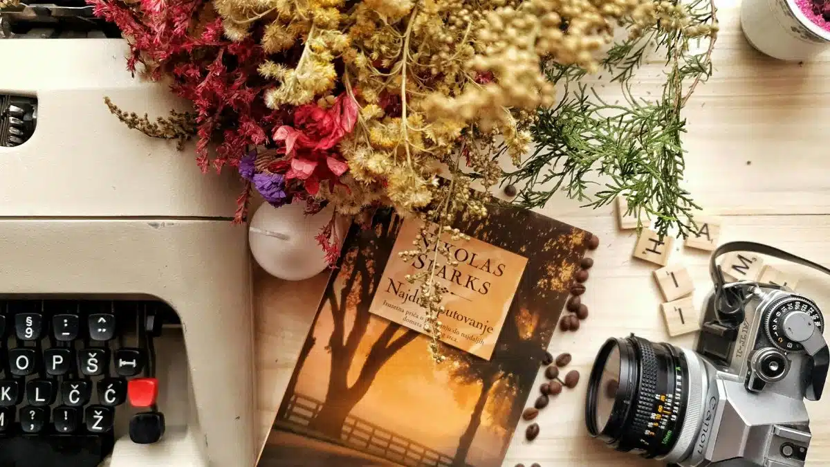 Top view of book and photo camera placed on wooden table near vintage typewriter composed with beautiful dried flowers