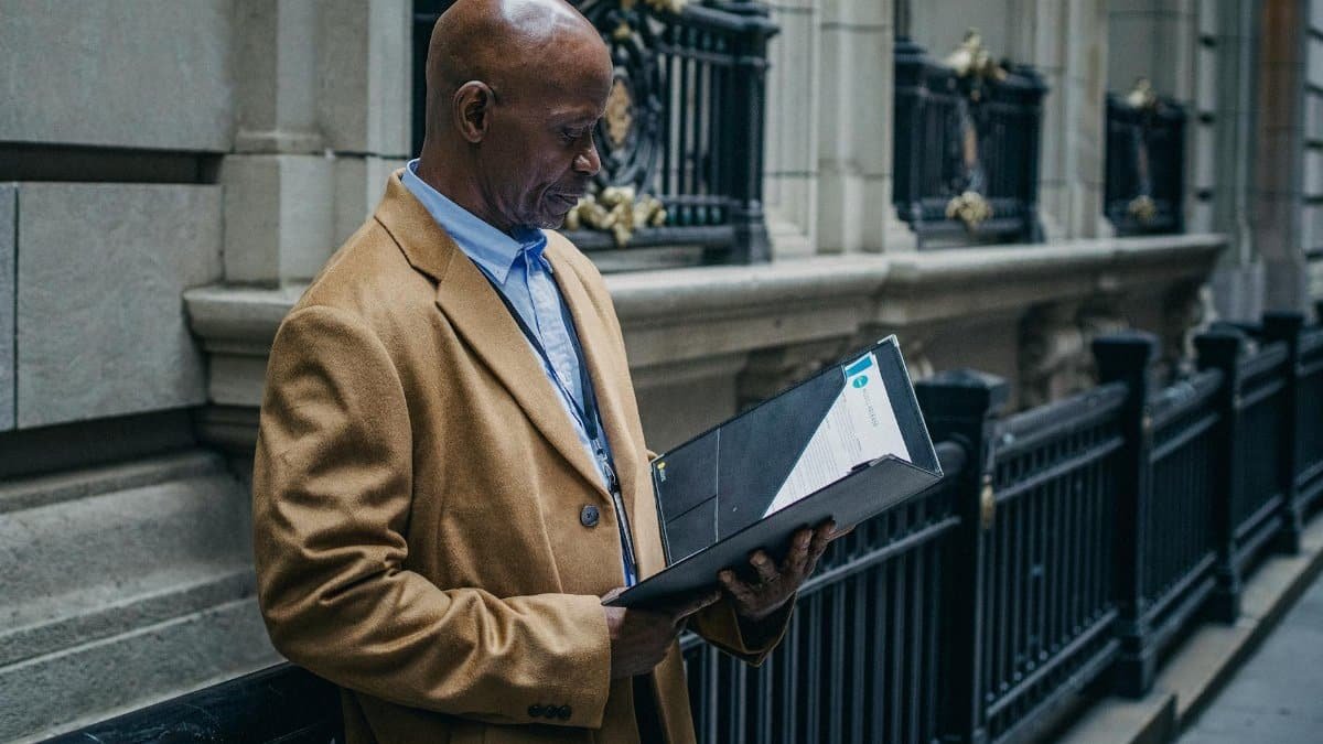 Side view of serious bald African American male examining papers in folder on street with metal fence