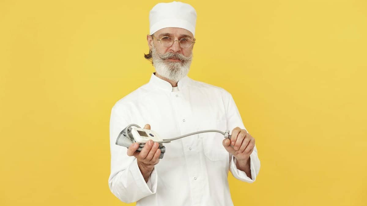 A senior chef in white attire holding a kitchen blender, standing against a yellow background.