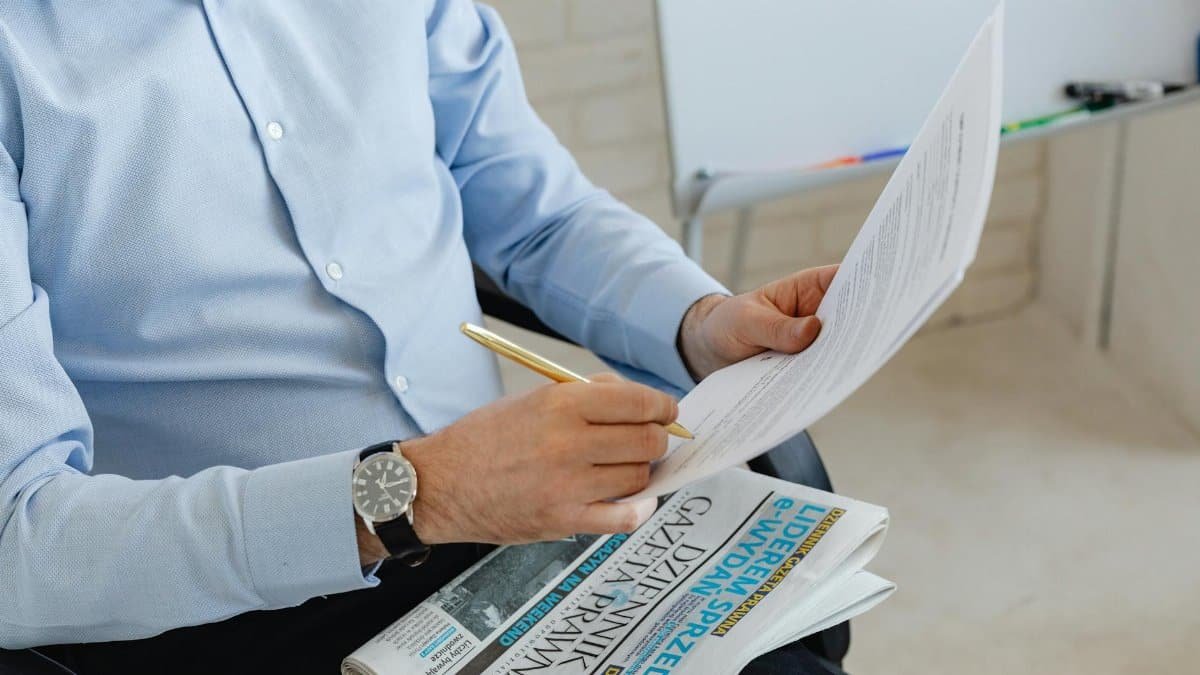 Businessman in blue shirt reviewing documents with a pen over a newspaper in an office setting.