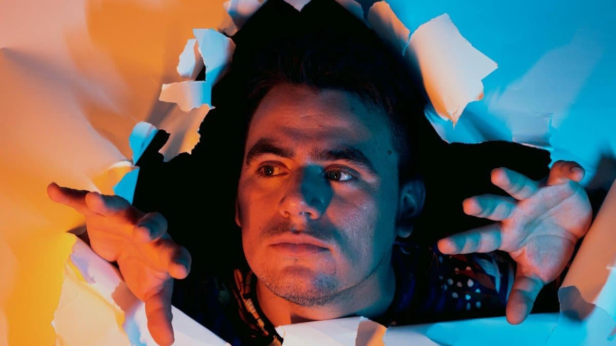 A man in vibrant lighting emerges through torn paper, symbolizing breakthrough and creativity.