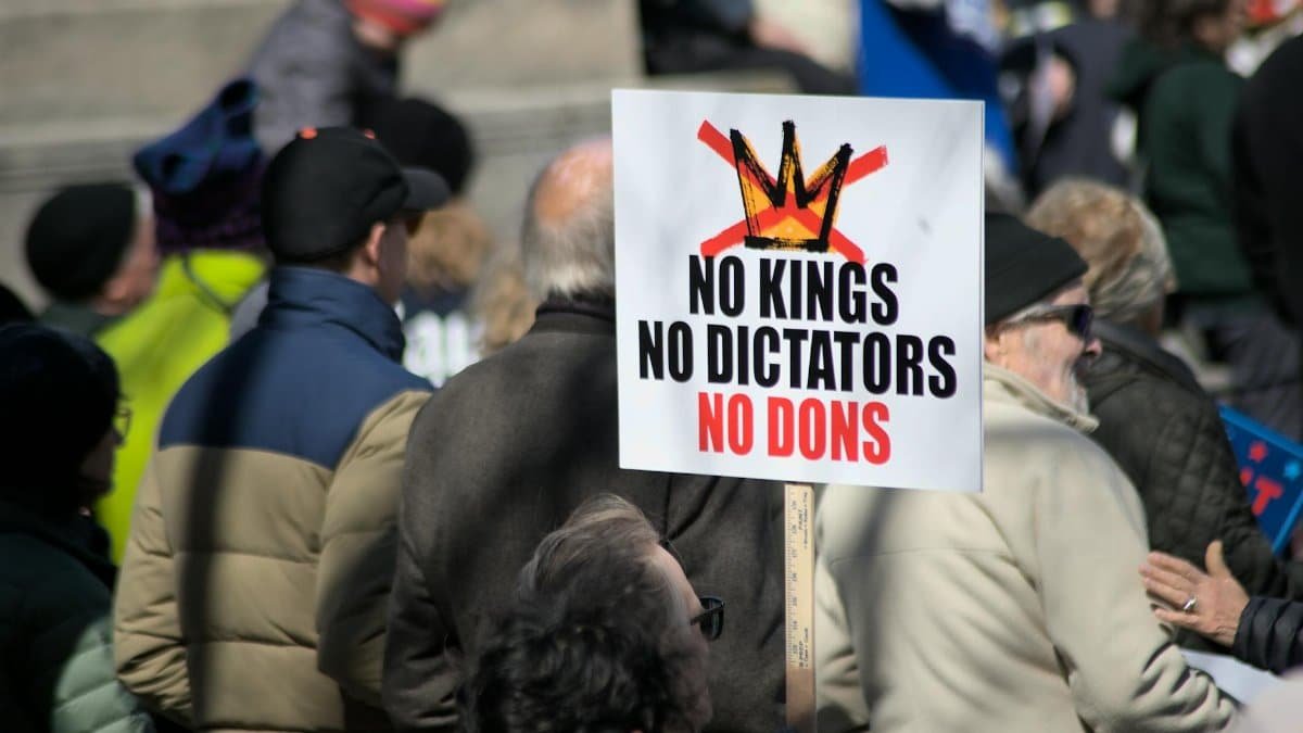 A crowd in Baltimore holds signs protesting against kings, dictators, and dons, promoting democracy.