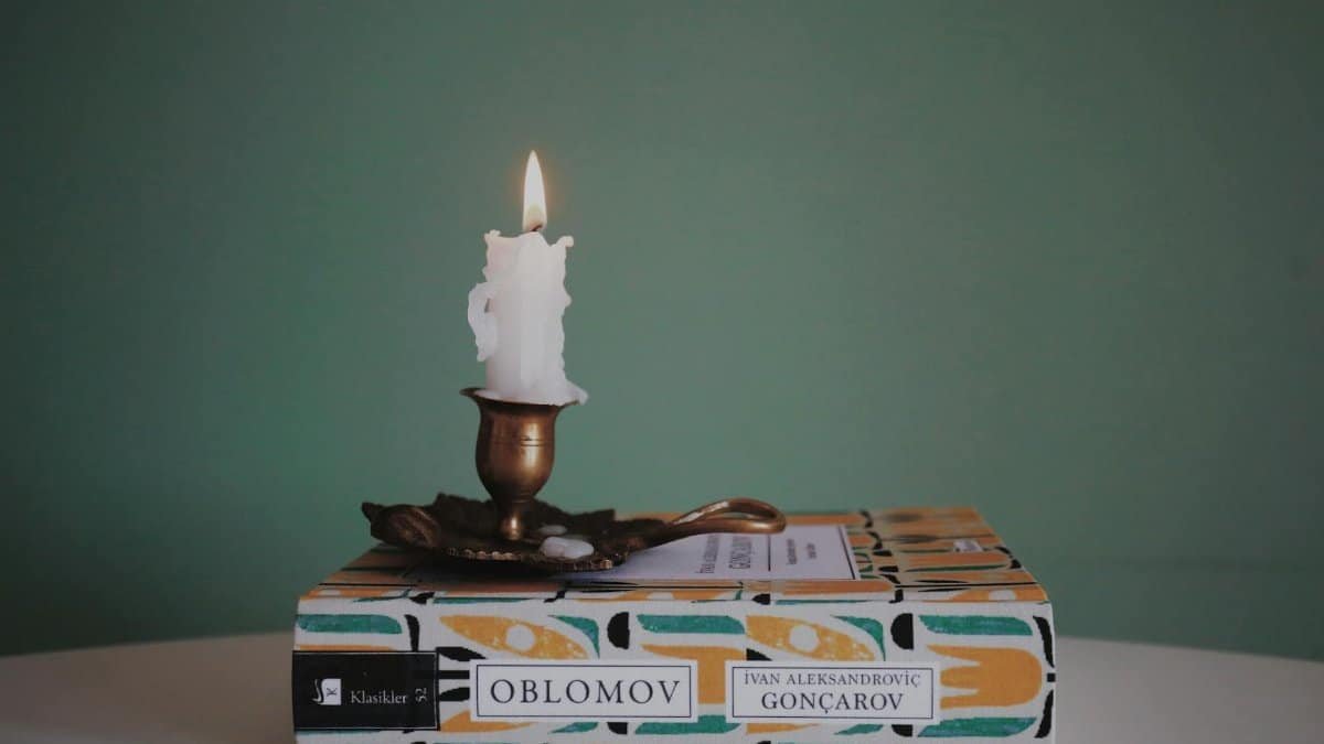 A lit wax candle on a brass holder rests atop the book Oblomov against a green backdrop.