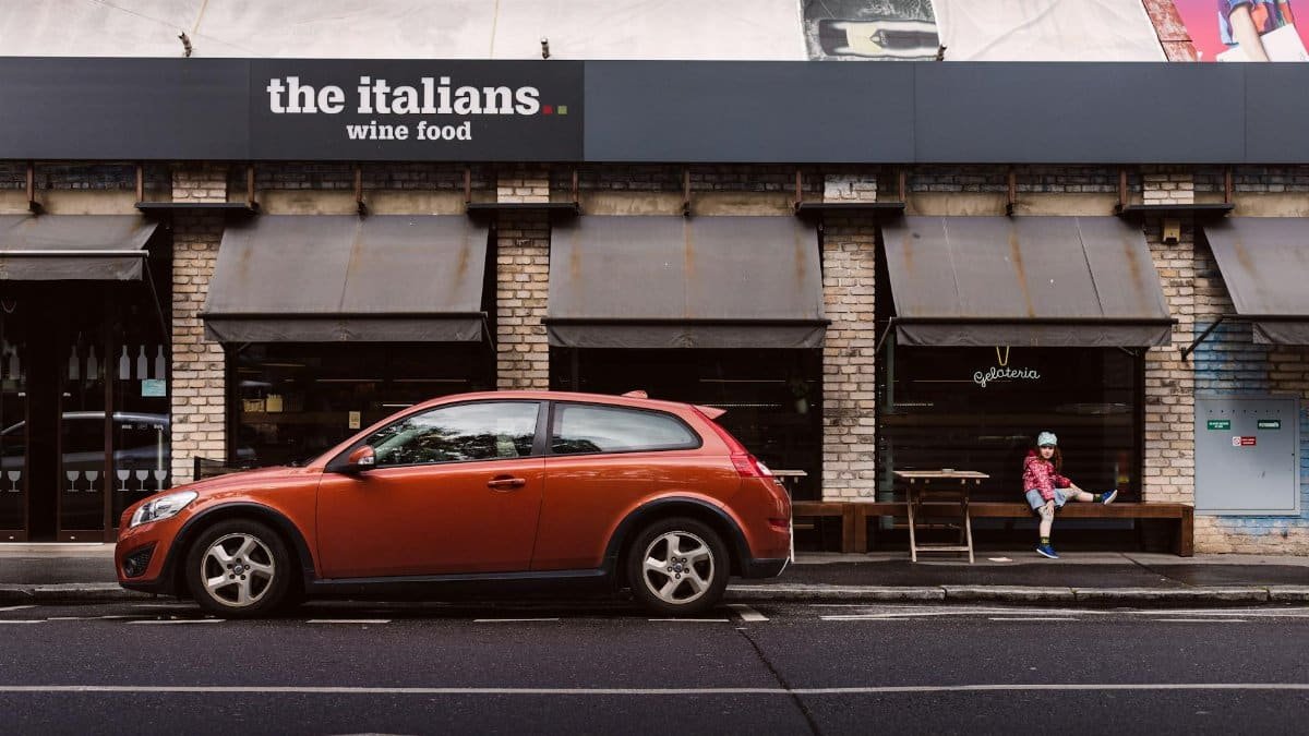 Red car parked outside a gelateria and Italian restaurant in Prague, with a child sitting nearby.