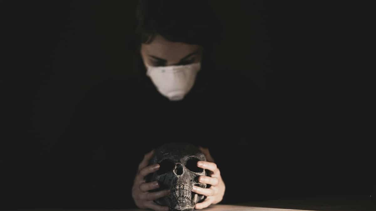A person wearing a mask holds a skull under a spotlight in a dark studio, symbolizing mortality and protection.