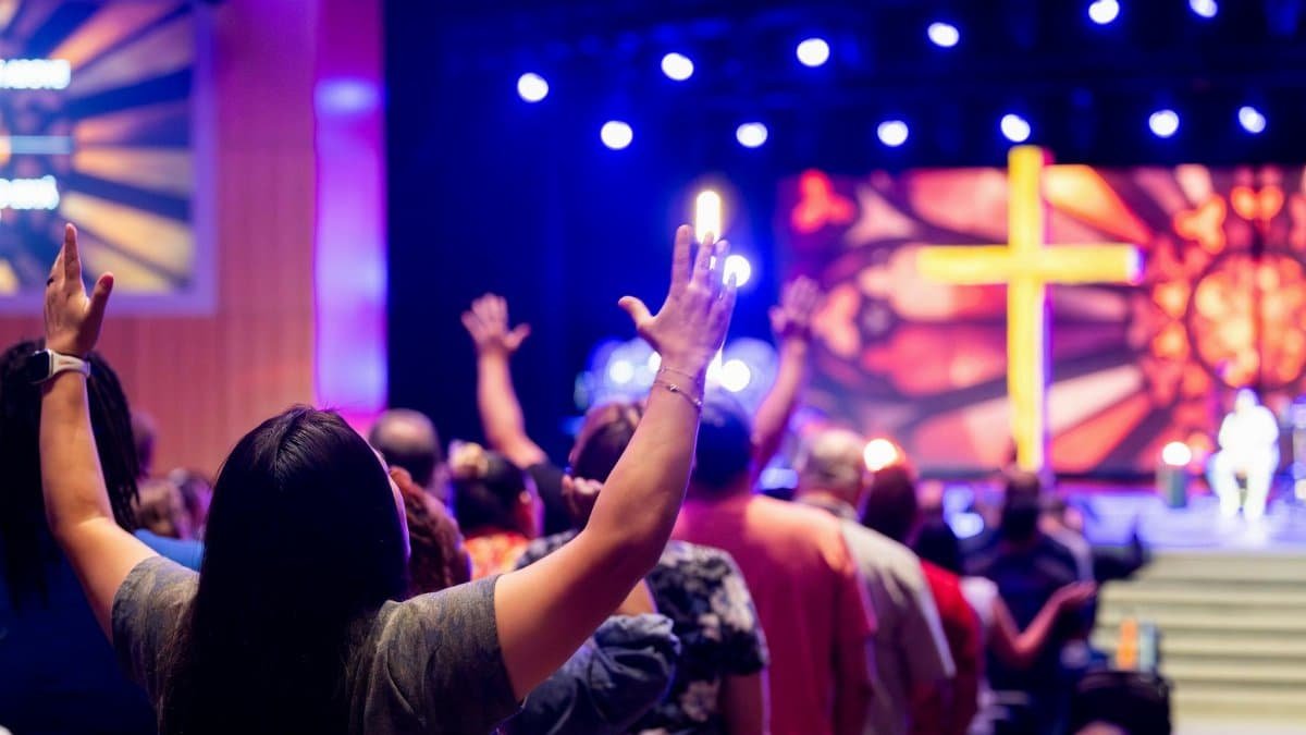 People engaged in a vibrant worship service, raising hands in faith under colorful stage lights and a prominent cross.