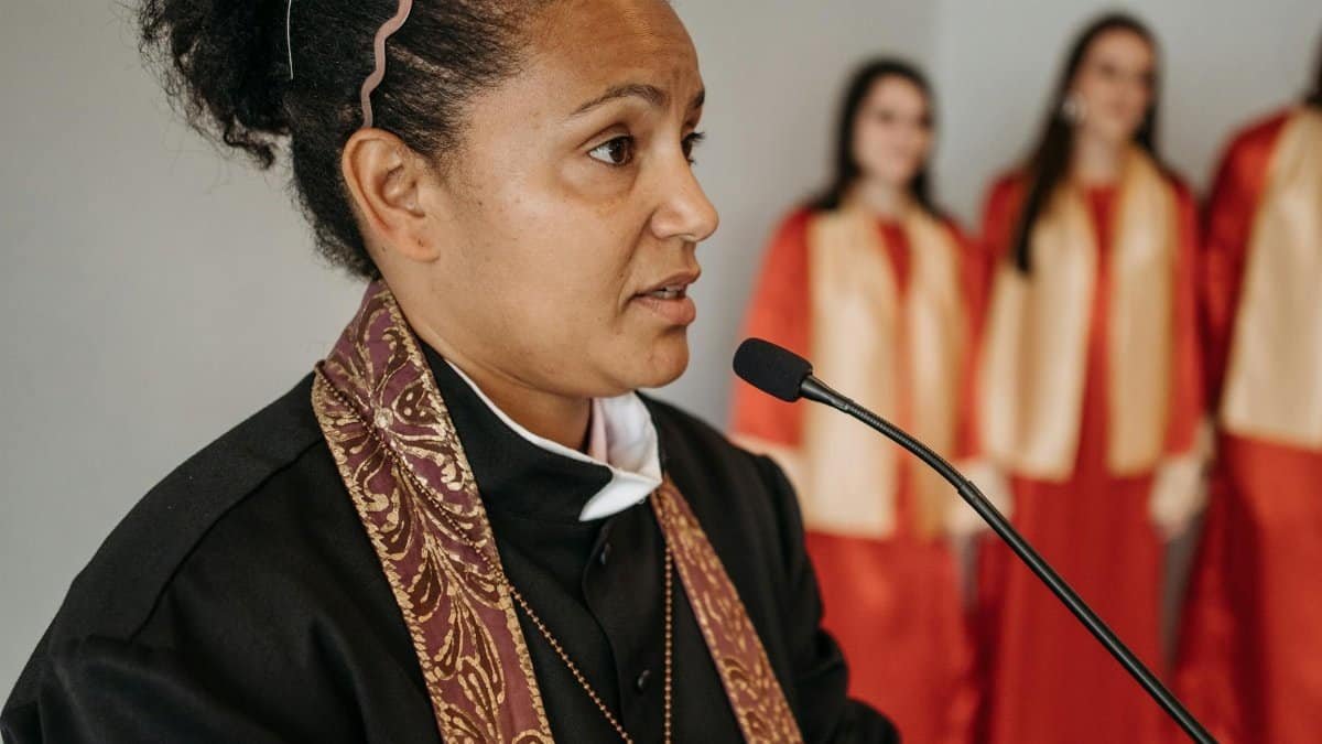 An African American woman pastor speaks at a church with a choir in the background, highlighting diversity and faith.