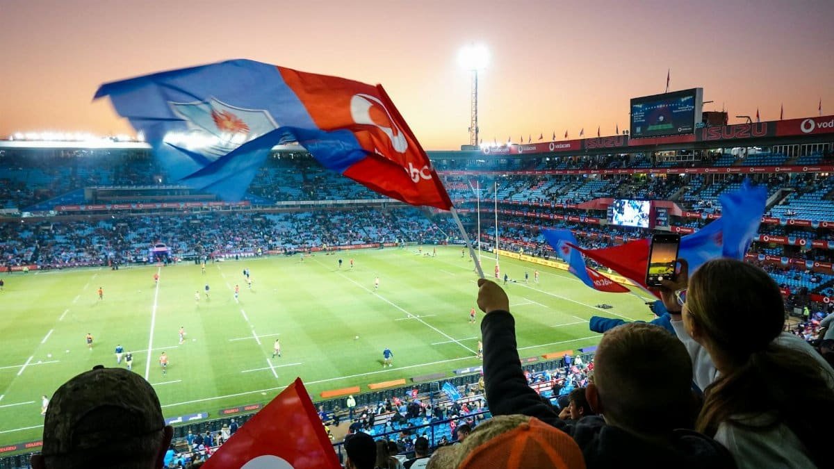 Fans cheer with flags in a large stadium during a rugby match at sunset, creating a lively atmosphere.