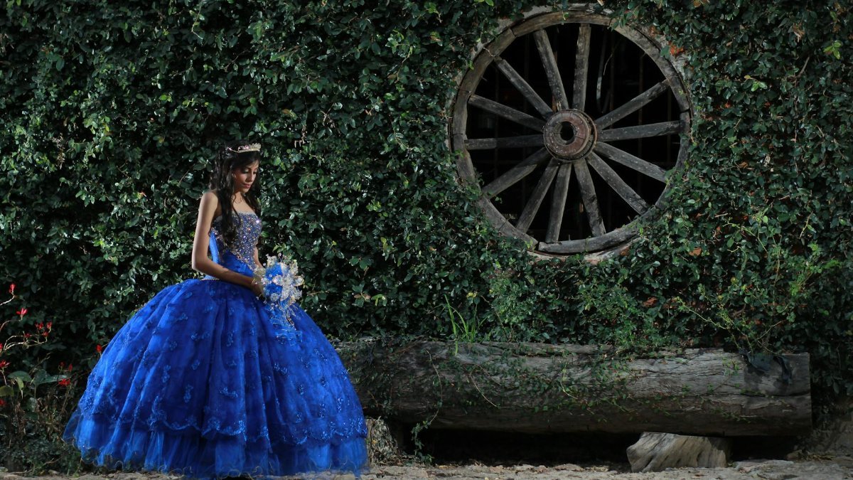 Young girl in a blue gown posing elegantly by a vintage wheel.