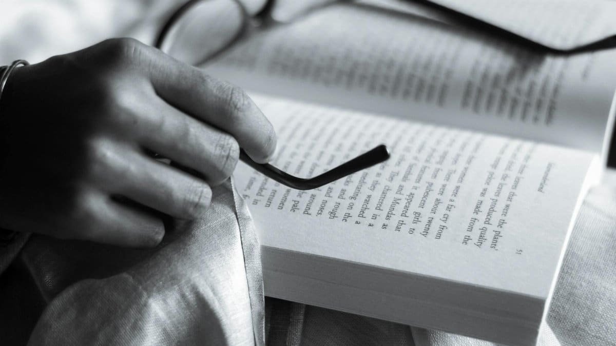 A detailed black and white close-up of a hand holding eyeglasses on an open book, creating an intellectual ambiance.