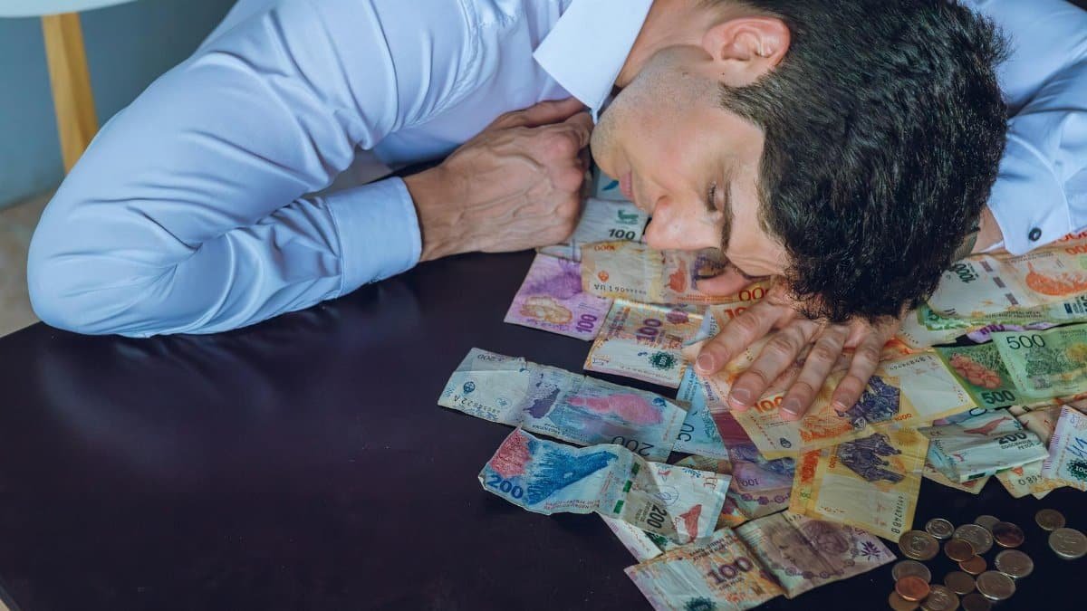 A tired man rests on a table covered with Argentine pesos, symbolizing financial stress.