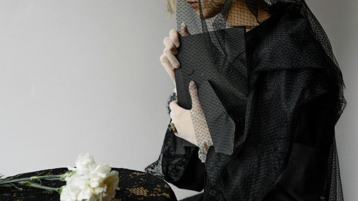 A woman dressed in black mourning attire holds a picture frame beside white flowers.