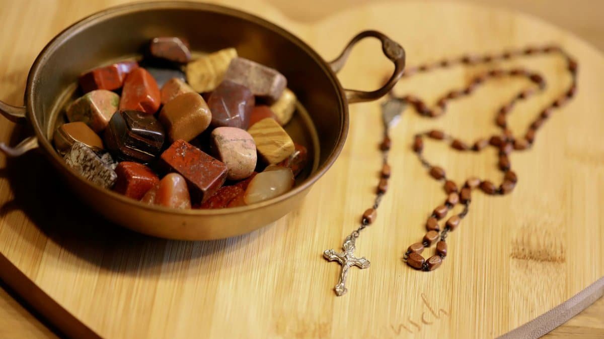 Colorful healing stones in bowl alongside rosary on wooden board symbolizing spirituality and tranquility.