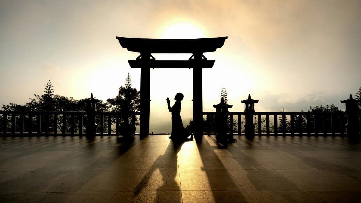 Silhouette of a person kneeling in prayer under a Torii gate at sunset, evoking peace and reflection.