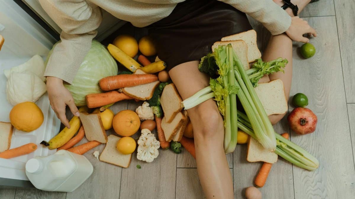 A woman amidst spilled groceries, highlighting food waste and consumerism with vegetables, fruits, and bread.
