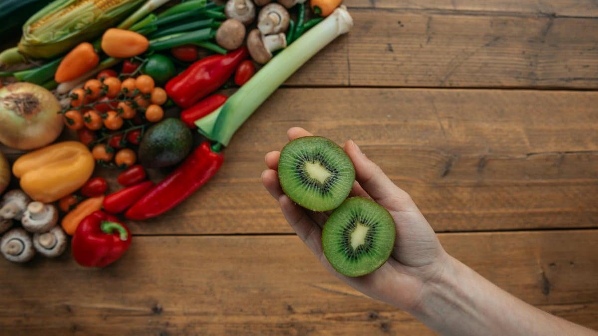 A hand holds sliced kiwi above a wooden table with fresh vegetables, highlighting healthy ingredients.