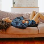 A woman in casual wear experiences abdominal discomfort while lying on a pink couch indoors.