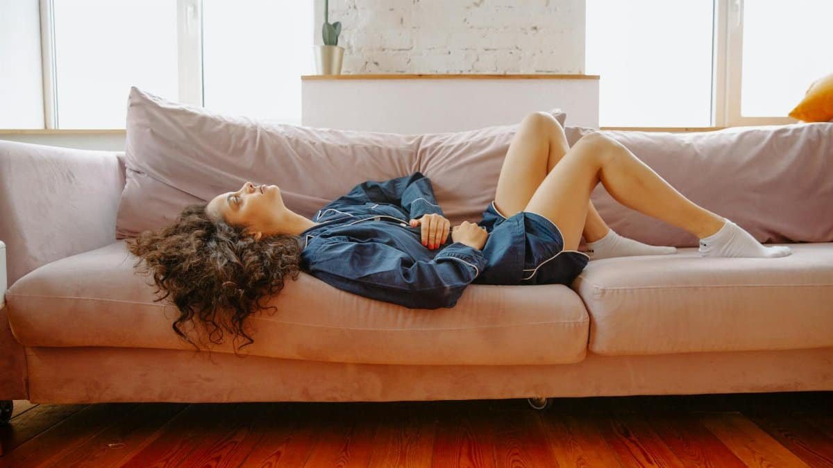 A woman in casual wear experiences abdominal discomfort while lying on a pink couch indoors.