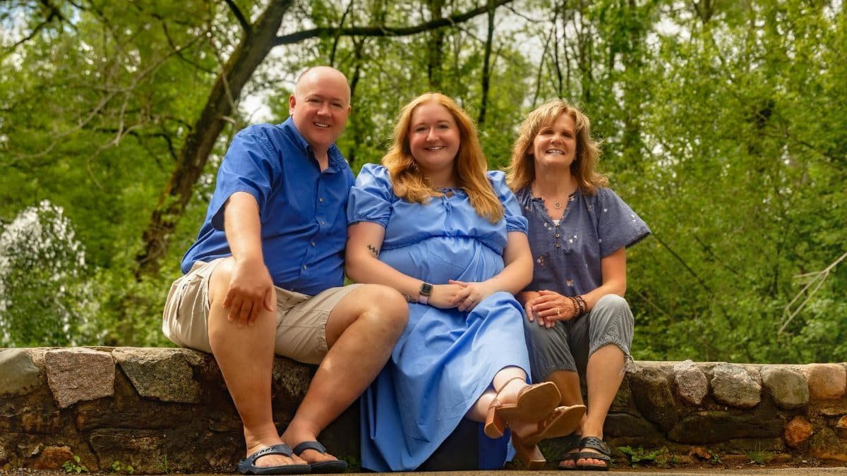 A joyful family of three sitting on a stone bench in a lush green outdoor setting.