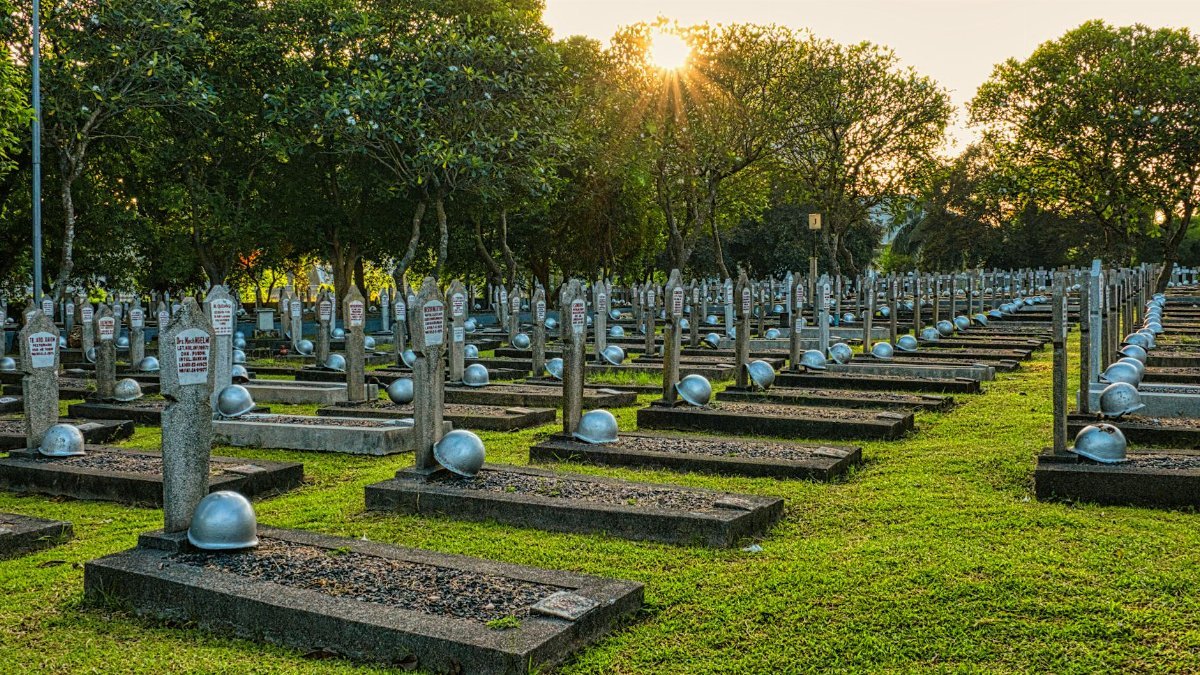 Rows of tombstones with military helmets located on grassy ground near tall lush green trees in heroes cemetery in Kalibata
