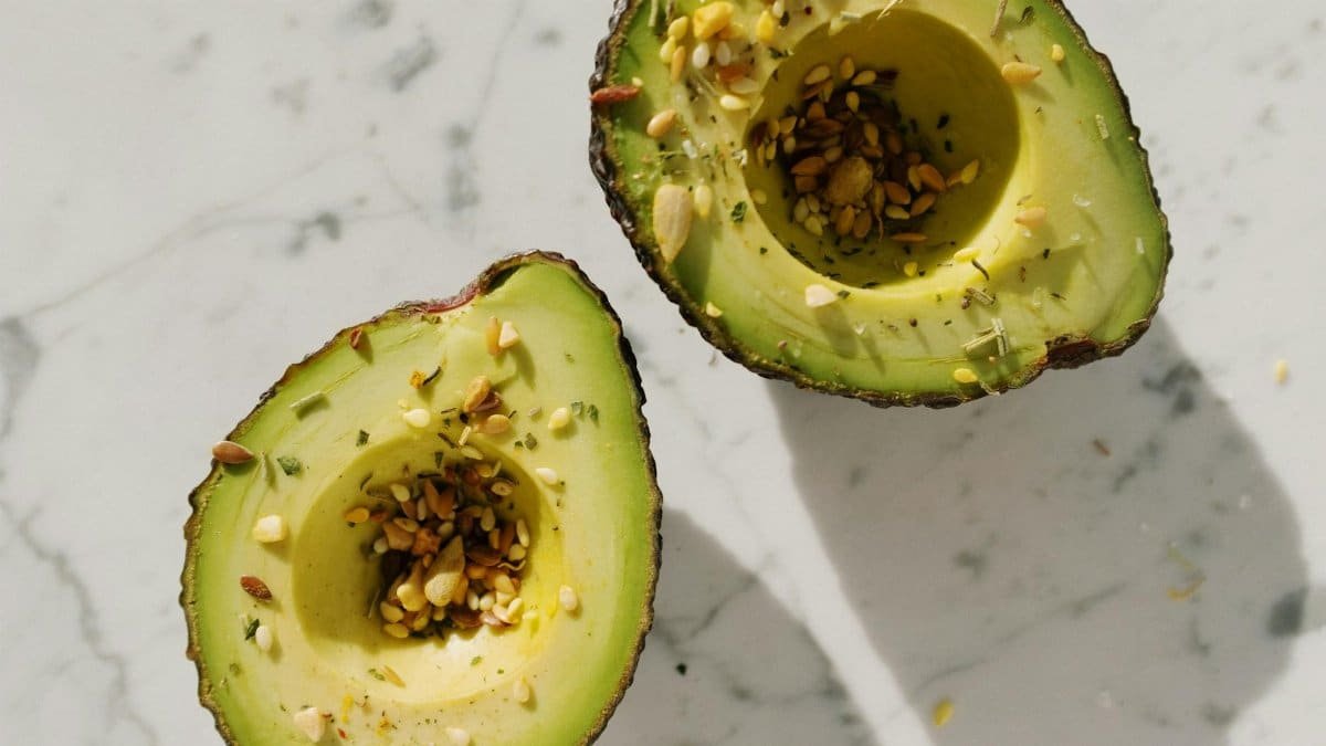 Top view of fresh green halved avocado without kernel with seed mixture on them placed on white marble table for healthy vegetarian breakfast