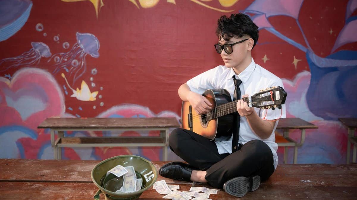 Young musician playing acoustic guitar indoors, busking next to a helmet with money.